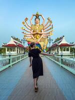 A woman walks along a bridge towards the grand, multi-armed statue of the Goddess Guanyin in Thailand. The statue's vibrant colors contrast with the clear blue sky, creating a striking visual photo