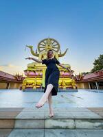 A young woman in a black dress joyfully poses barefoot on temple steps, aligning her arms with the golden Big Buddha statue at sunset. Shot from a low angle for a dramatic effect photo