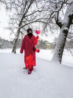Smiling in a snowy park, a woman in a pink geometric-patterned coat and matching boots has a pixelated heart hovering above her hand. photo