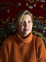 A woman clad in a comfortable orange sweater is seated on a patterned sofa in a traditional Russian dwelling, a lavishly ornamented vintage rug adorning the wall behind her. photo