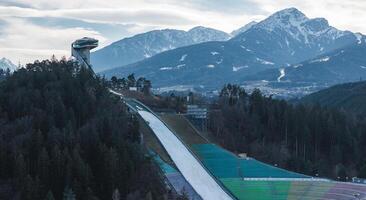 The Bergisel Ski Jump in Innsbruck, Austria, with its modern tower, steep ramp, colorful seating, and snow capped Alpine mountains in the background. photo