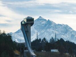 The Bergisel Ski Jump in Innsbruck, Austria, features sleek modern architecture, surrounded by trees, buildings, and alpine peaks in the background. photo