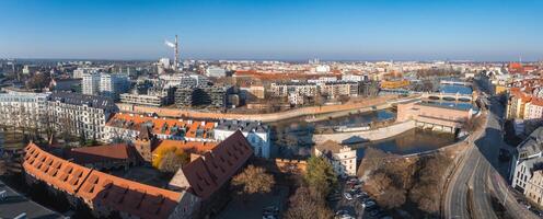 Aerial view of Wroclaw, Poland, featuring red roofed historic buildings, modern structures, the Oder River, arched bridges, and a tall smokestack. photo