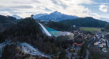 Aerial view of the Bergisel Ski Jump in Innsbruck, Austria, surrounded by forests, residential areas, and open fields with the Alps in the background. photo