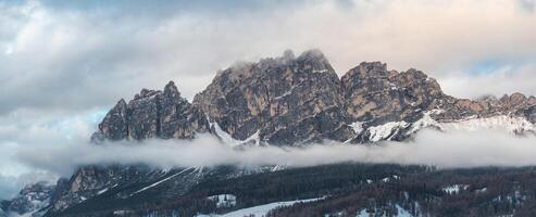 Aerial view of rugged Dolomite peaks near Cortina d'Ampezzo, Italy, with snow covered slopes, low clouds, and visible ski trails and structures. photo