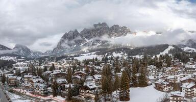 aéreo ver de cortina d'ampezzo en Italia con nieve cubierto tejados, alpino edificios, hojas perennes árboles, y dolomitas picos parcialmente en nubes foto