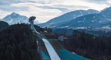 The Bergisel Ski Jump in Innsbruck, Austria, stands on a forested hill with its modern design, surrounded by dense forests and alpine mountain ranges. photo