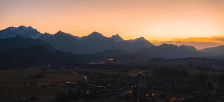 Neuschwanstein Castle glows at sunset, surrounded by dark hills, the Bavarian Alps, and a village with scattered lights in the foreground. photo