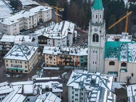 aéreo ver de cortina d'ampezzo en el dolomitas, presentando el basílica menor campana torre, nieve cubierto tejados, y construcción grúas en el antecedentes. foto