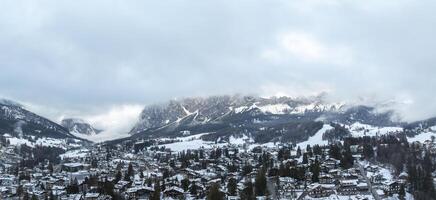 Aerial view of Cortina d'Ampezzo, Italy, with snow covered rooftops, alpine architecture, and the Dolomites partially shrouded in clouds. photo
