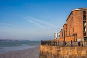 Historic Liverpool waterfront featuring a red brick Albert Dock building, curved design, iron railings, and the River Mersey under a clear blue sky. photo