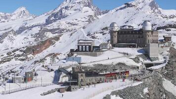 Aerial view of Gornergrat observatory with twin domes, Matterhorn peak, red train arriving, and skiers moving on snowy slopes in the Swiss Alps. video