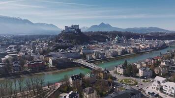 Aerial pan of Salzburg, Austria, featuring Hohensalzburg Fortress, the Salzach River, historic architecture, and snow capped mountains in the background. video