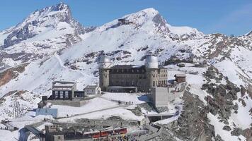 Smooth aerial pan of Gornergrat observatory with Matterhorn in the background, surrounded by snowy peaks, ski trails, and a clear blue sky. video