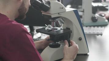 Medical worker looks at blood sample through a microscope, makes analysis in laboratory. Laboratory assistant conducts histological research of biomaterial using modern microscope video