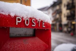 A red postal box labeled 'POSTE' is partially covered in snow, set against a blurred view of buildings with balconies in a snowy town, likely Cortina. photo