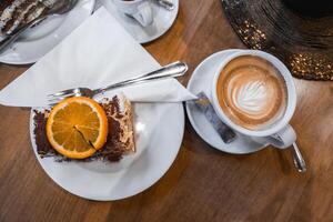 A slice of tiramisu with orange garnish on a white plate, paired with a cappuccino featuring latte art, set on a wooden table in a luxury setting. photo