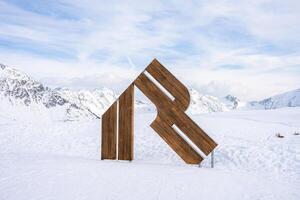 A wooden sculpture resembling an abstract letter R stands in snowy terrain, surrounded by rugged alpine mountains under a clear blue sky with clouds. photo