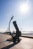 A large telescope like sculpture on a paved promenade in Liverpool, with the River Mersey, industrial structures, and a tall chimney in the background. photo