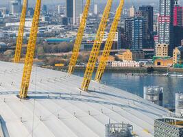 Close up aerial view of the O2 Arena in London with its white dome and yellow masts, the River Thames, and modern high rise buildings in the background. photo
