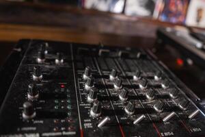A professional audio mixer with knobs, sliders, and buttons on a wooden surface in a dimly lit room. Blurred album covers are visible in the background. photo