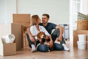 Front view. Sitting together. Young couple with dog are moving to new home photo