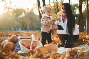 Beautiful mother is with her son in the autumn forest. Boy is holding orange leaf. Butternut squash and fruits on the ground photo