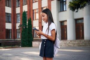 Wireless ,device, in hands, standing. School girl in uniform is outdoors near the building photo