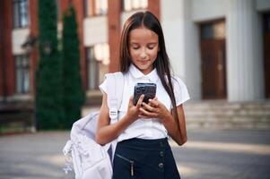 Holding ,smartphone. School girl ,in uniform is outdoors near the building photo