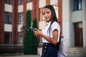 Holding smartphone. School girl, in ,uniform is outdoors near the building photo
