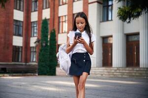 Phone in hands. School girl in ,uniform, is outdoors near the building photo