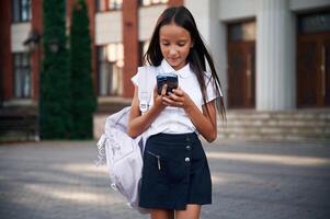 Phone in hands.. School ,girl in uniform is outdoors near the building photo
