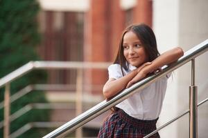 Leaning on the railing. School girl in uniform is outdoors near the building photo