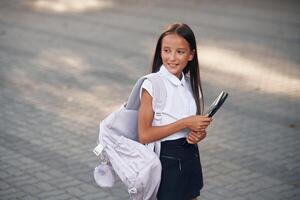 Schoolgirl with tablet and backpack is standing outdoors photo