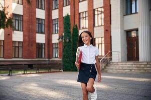 Standing, front view,, holding books., School girl in uniform is outdoors near the building photo