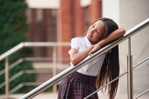 Leaning, on the ,railing. School girl in uniform is outdoors near the building photo