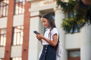 Using, smartphone. School girl in uniform ,is outdoors near the building photo