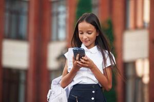 Using, smartphone. School, girl in uniform is outdoors near the building photo