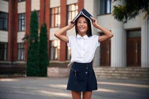 Playing ,with book that is over the head. School girl in uniform is outdoors near the building photo