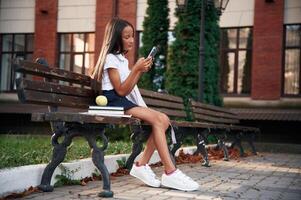 Sitting ,on ,the bench, with smartphone. School girl in uniform is outdoors near the building photo