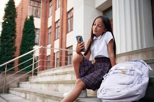 Sitting ,on the stairs, using, smartphone. School girl in uniform is outdoors near the building photo