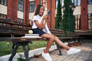 Sitting on, the bench with smartphone. School girl in uniform is outdoors near the building photo