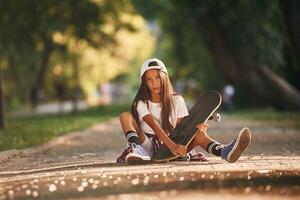 Front view, sitting on the ground.. Happy ,little girl with skateboard outdoors photo