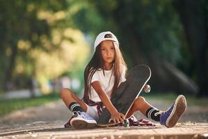 Front view, sitting on ,the ground. Happy little girl with skateboard outdoors photo