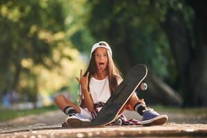 Front view, showing gestures,. Happy little girl with skateboard outdoors photo