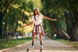 Green ,trees are in the background, on the path. Happy little girl with skateboard outdoors photo