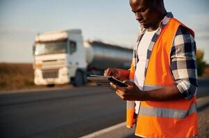 In orange colored uniform, with ,tablet.. Black man is standing on the road with truck on it photo