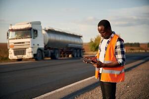In ,orange colored uniform, ,with ,tablet. Black man is standing on the road with truck on it photo