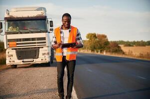 Front view. Black, man is standing on the road with truck on it photo