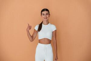 Showing thumb up, gesture. Beautiful young woman in white clothes is standing indoors photo